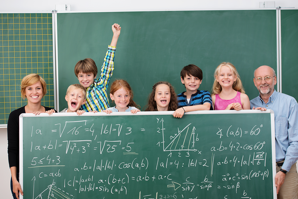 Eine Lehrerin und ein Lehrer stehen mit den Grundschülern vor der Schultafel und halten selbst auch eine große Tafel vor sich mit komplizierten mathematischen Ausdrücken.